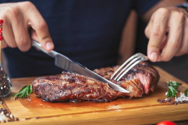 hands holding fork and knife and eating delicious juicy steak on a wooden plate